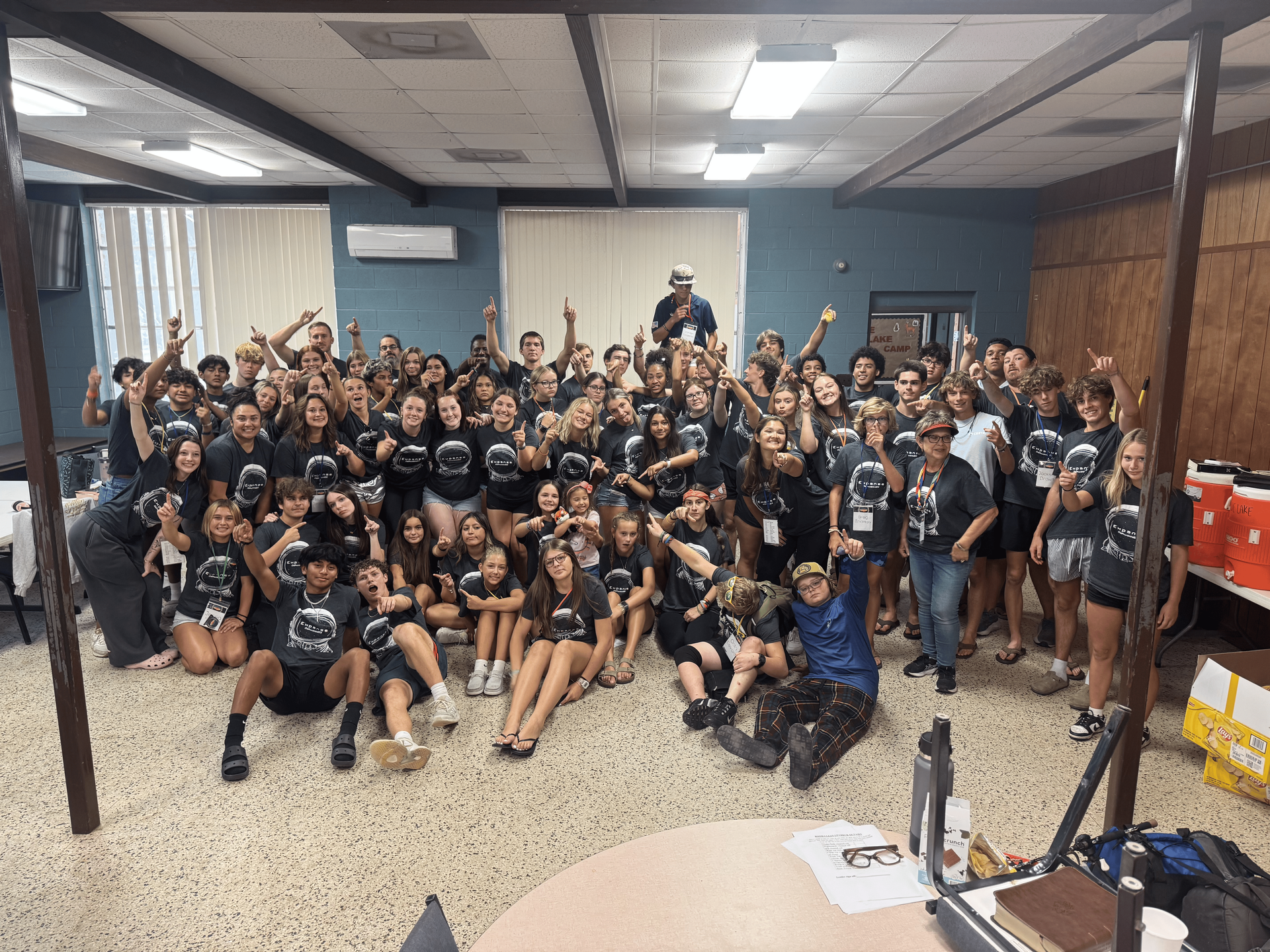 Large group posing indoors in a classroom, many wearing black shirts, with a speaker centered at the back.