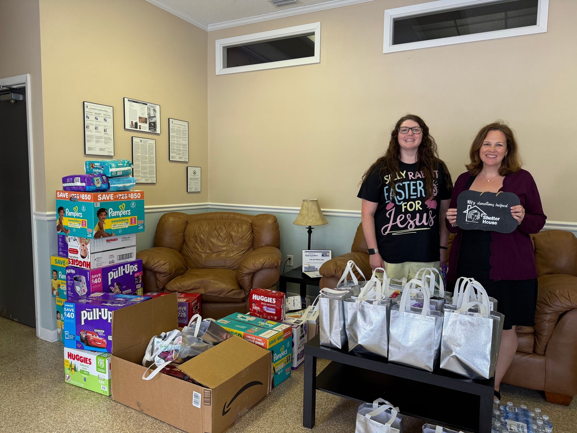 Two women stand beside stacks of donated supplies and gift bags in a living room.