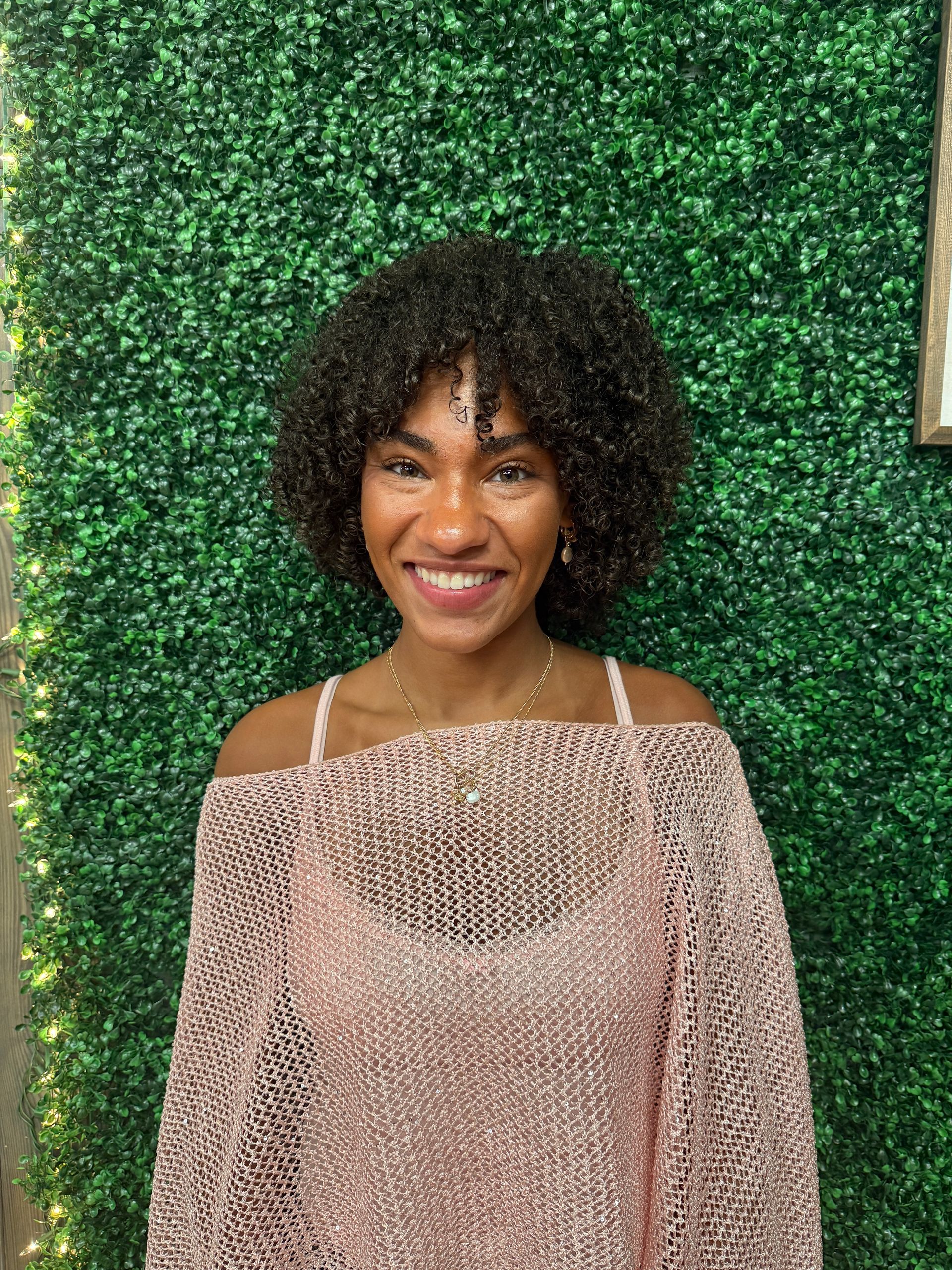 Woman smiling in front of a green textured wall, wearing a light pink off-the-shoulder top.