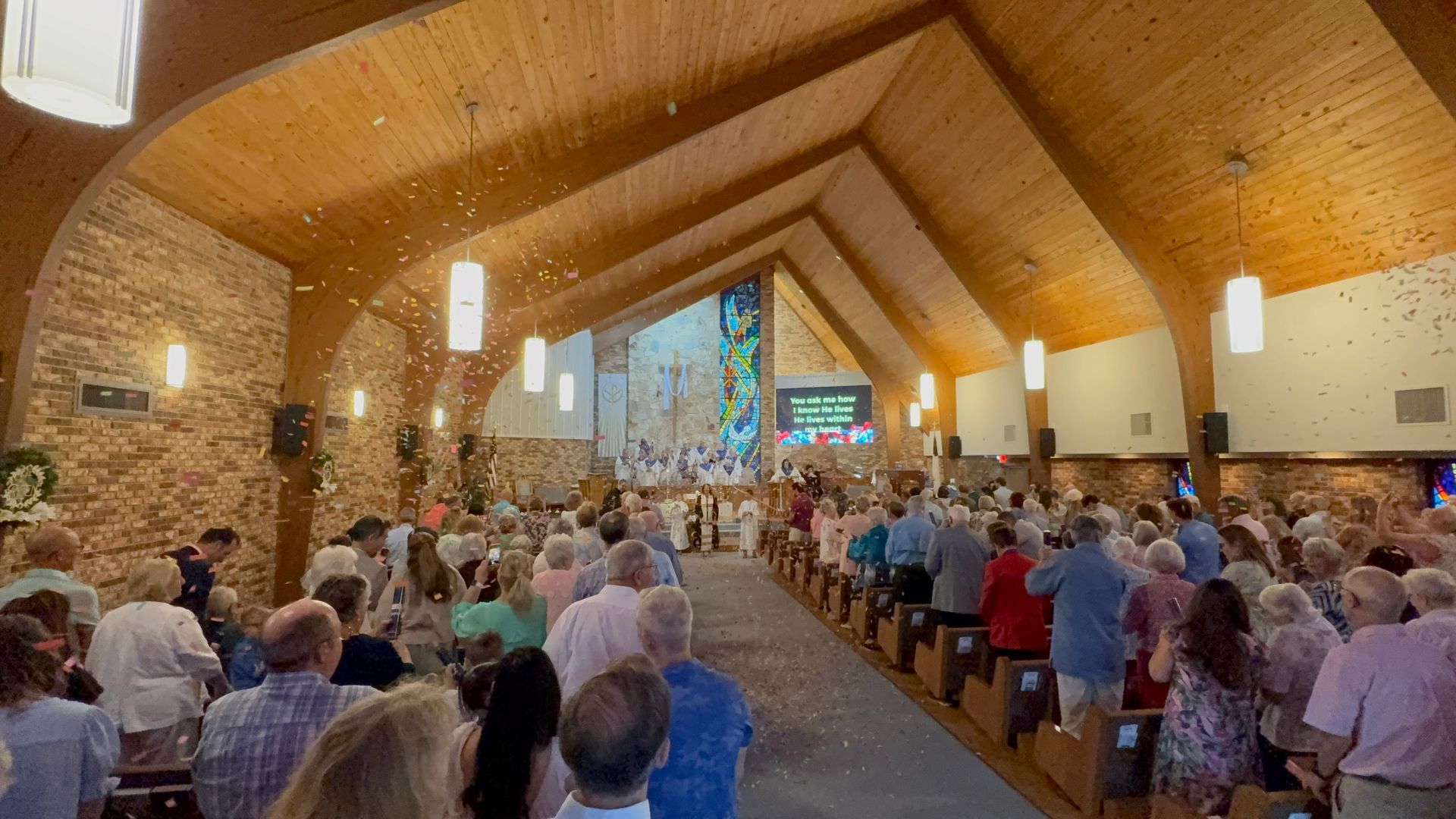 Crowded church service in a wooden sanctuary, people seated facing the altar with stained-glass windows at the front