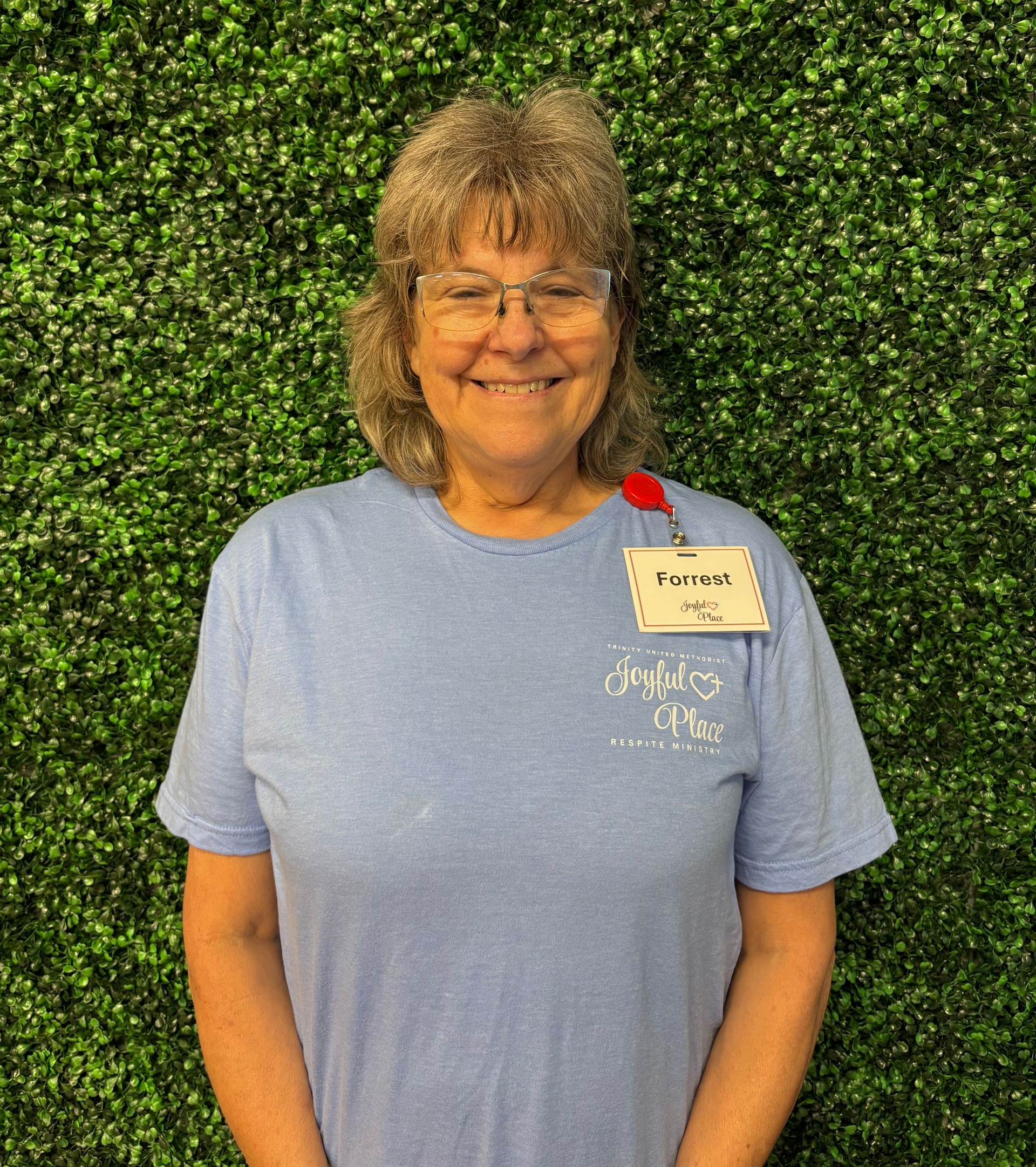 Smiling person in a light blue shirt with name tag standing before a green hedge backdrop