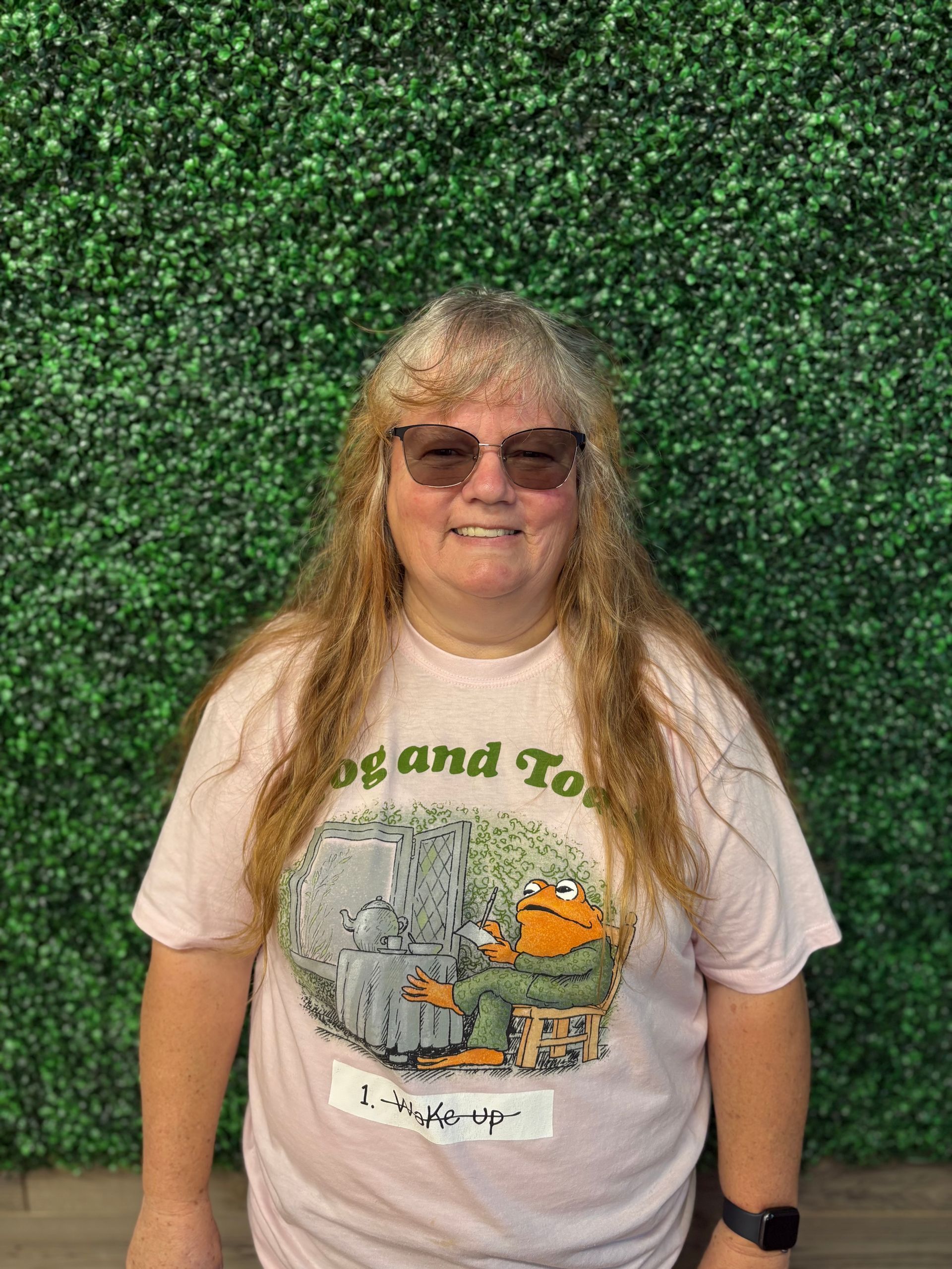 Person with long blonde hair and sunglasses, wearing a white graphic T-shirt, standing before a green hedge wall.