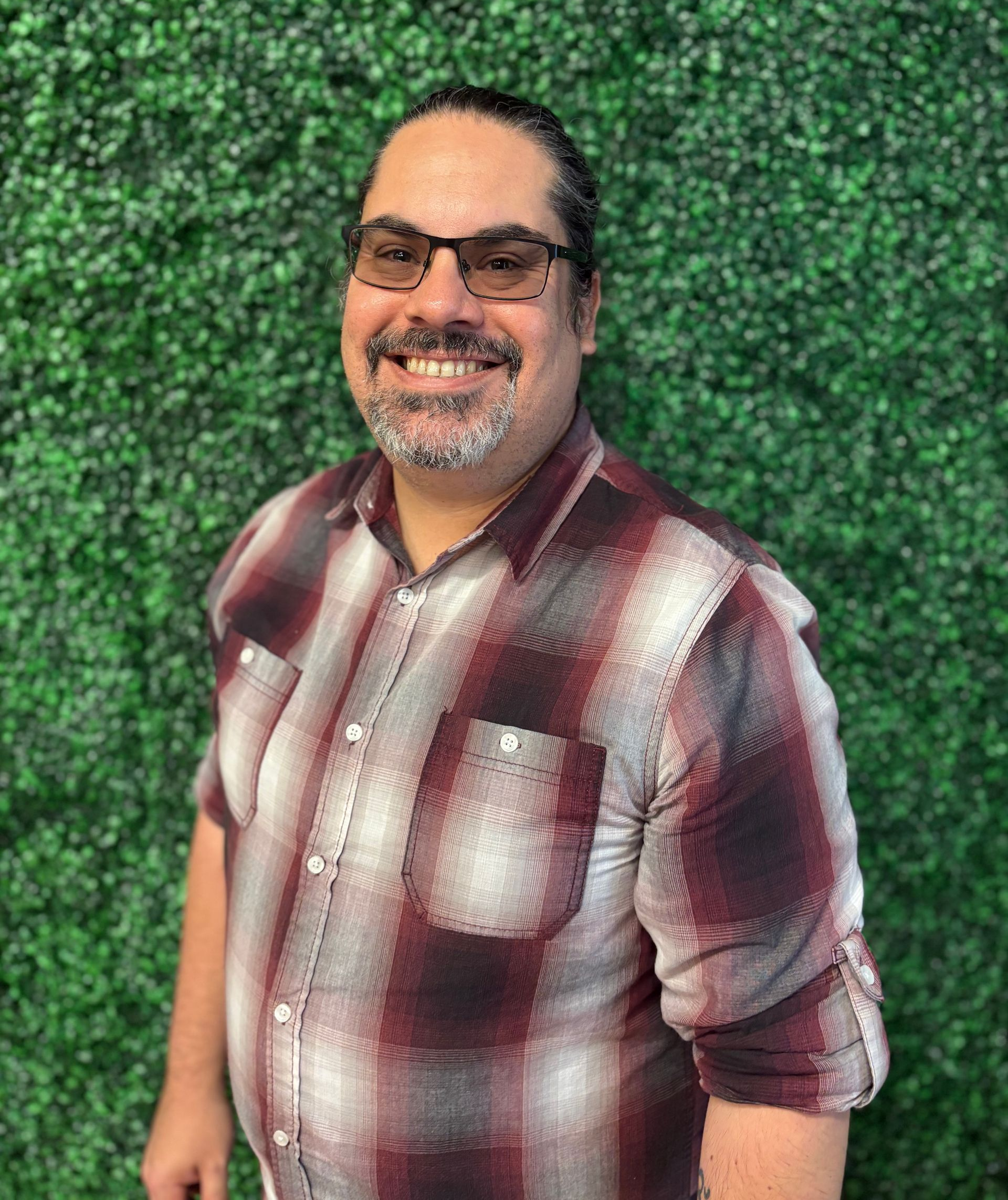 Smiling man in plaid shirt standing before a green leafy wall