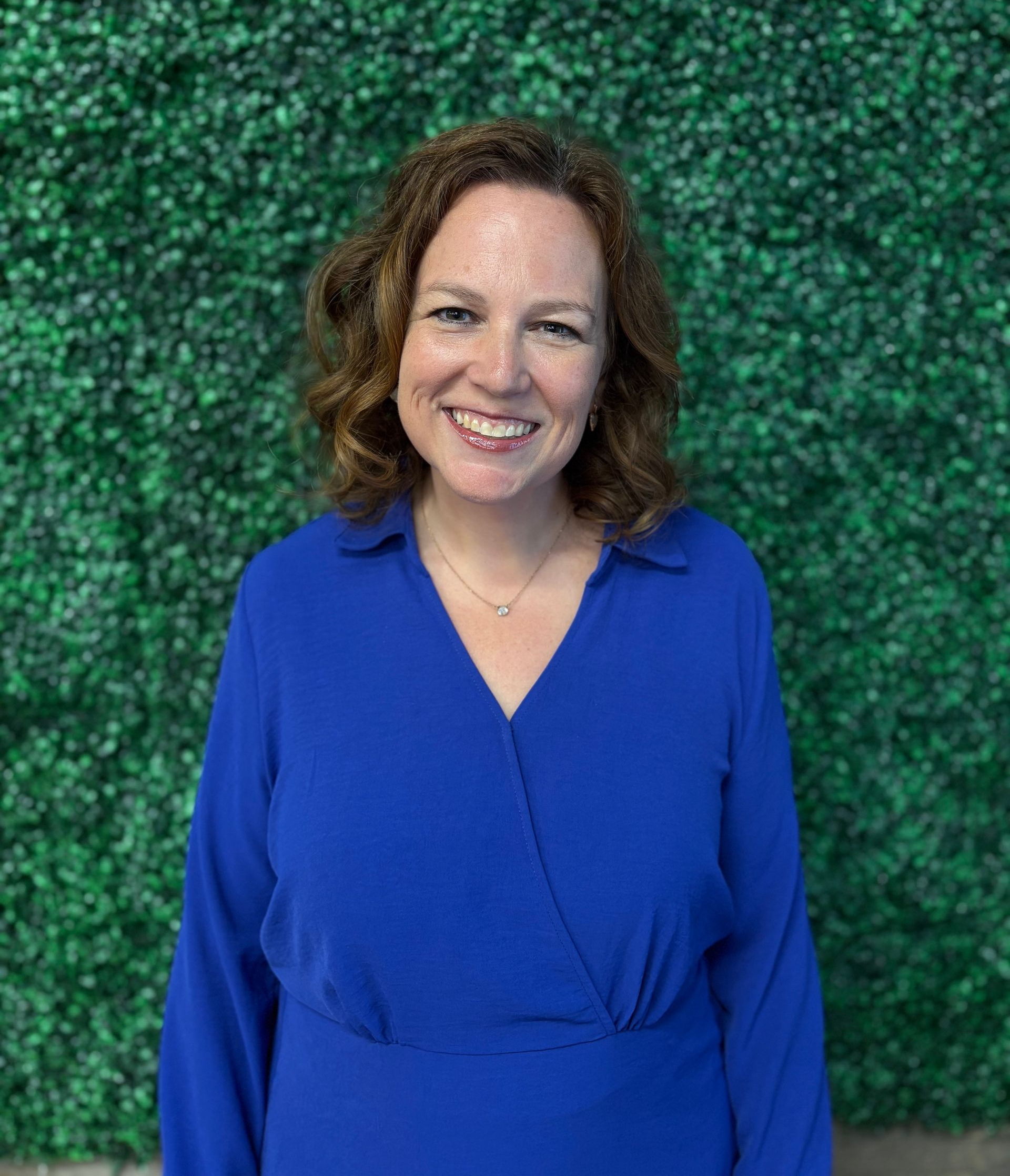 Smiling woman in a blue blouse standing before a green leafy wall background