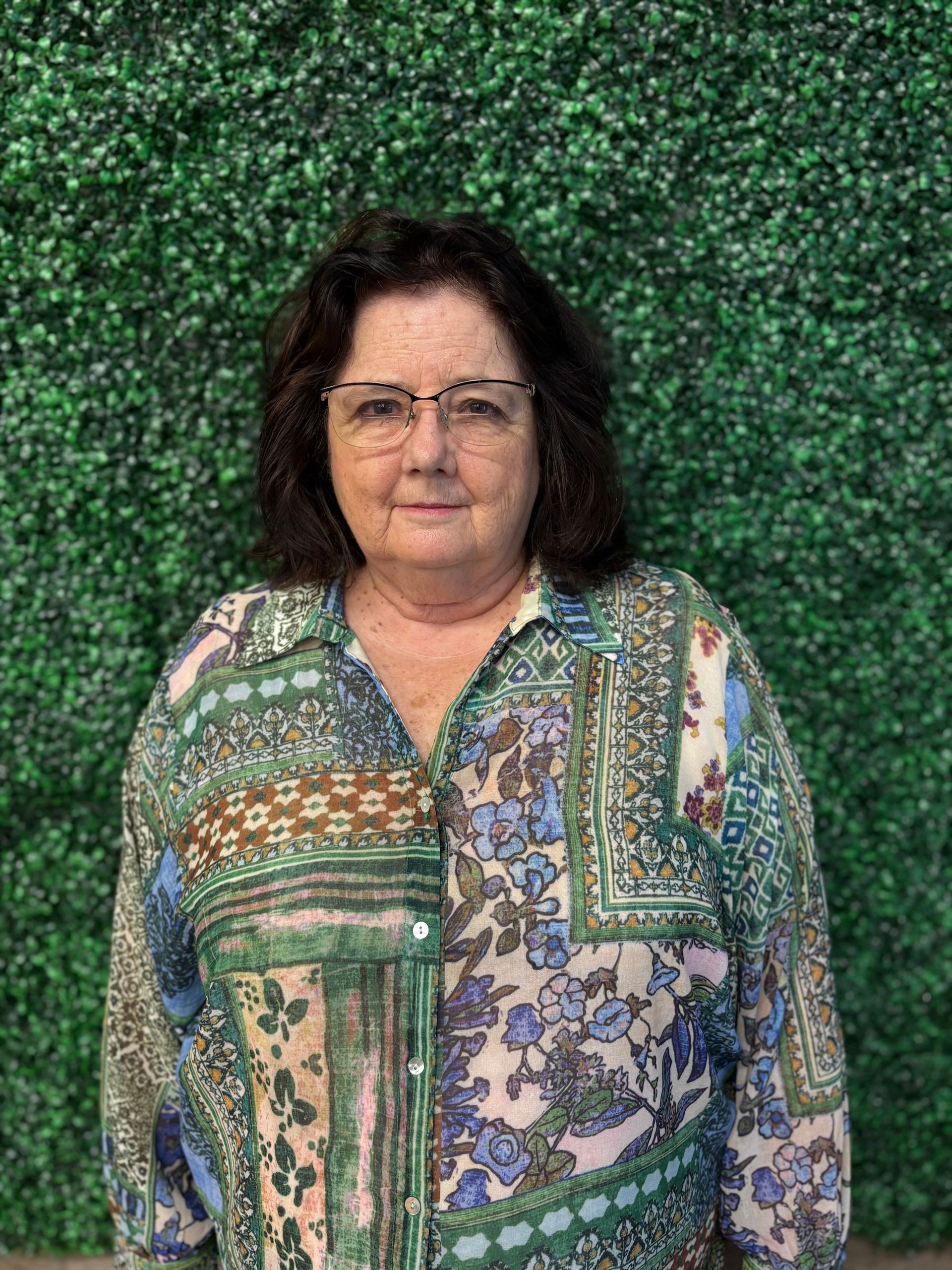 Woman in a patterned green and blue blouse standing before a leafy green backdrop