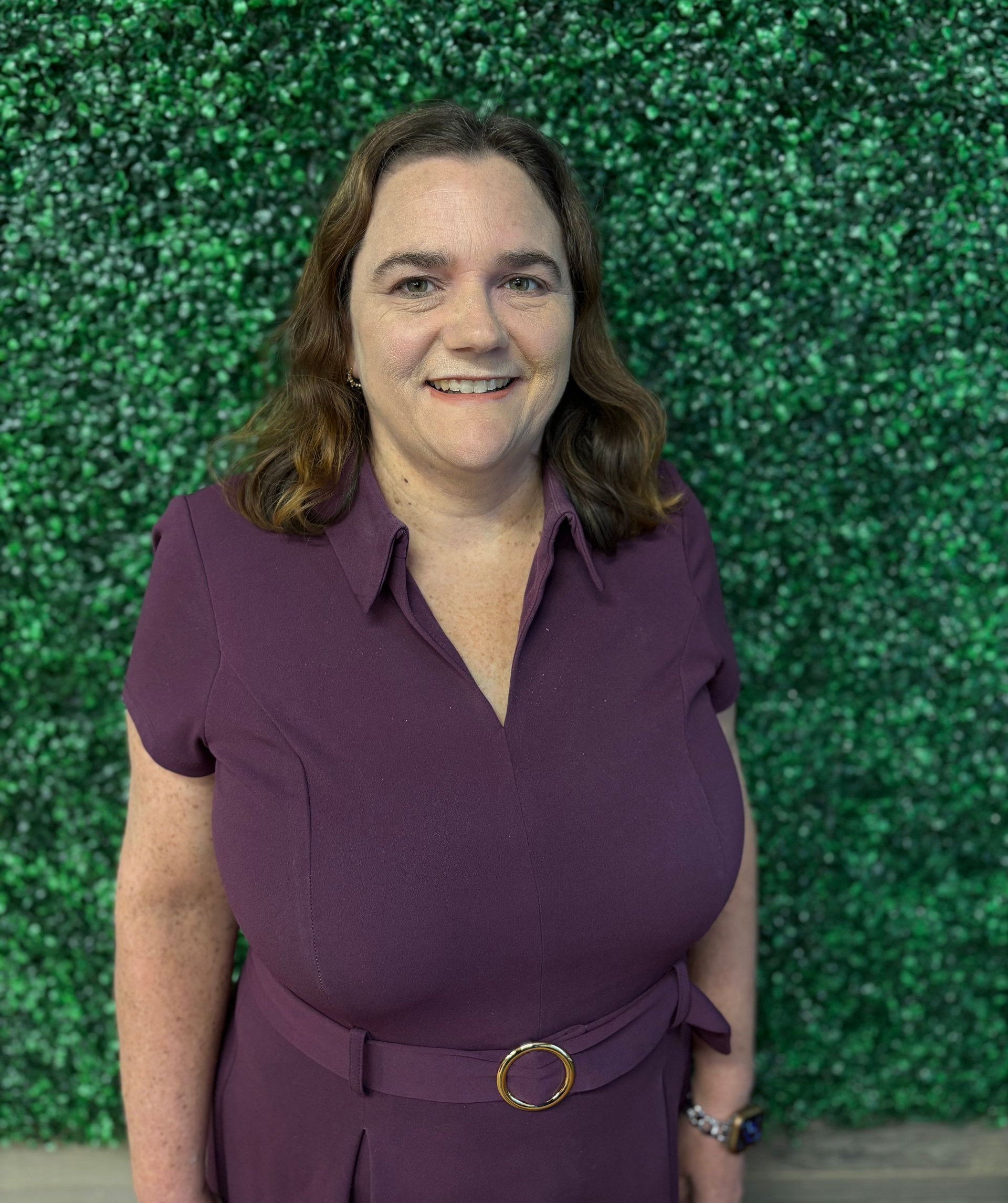 Smiling woman in a purple dress standing against a green leafy wall