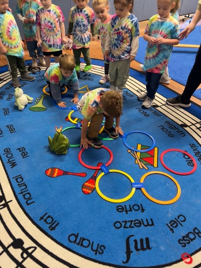 Children playing with Olympic rings on a classroom floor mat, surrounded by colorful props and a blue gym floor