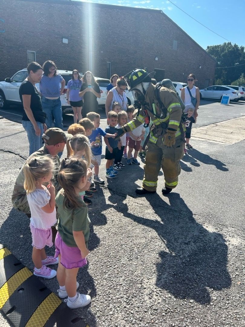 Firefighter in full gear speaking to a line of children outdoors in a parking lot