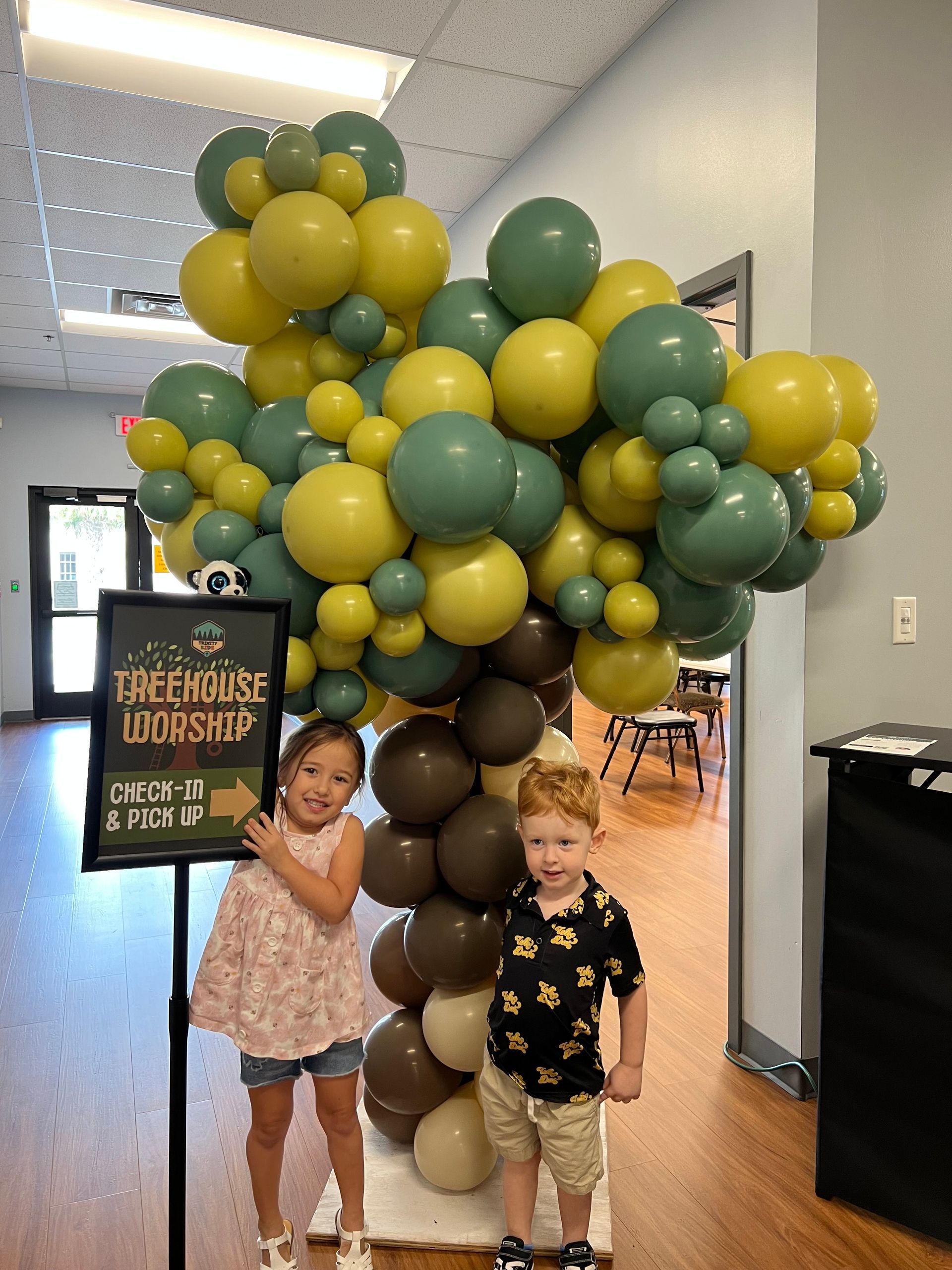 Two children standing by a large green, gold, and white balloon display in a hallway, holding a sign.