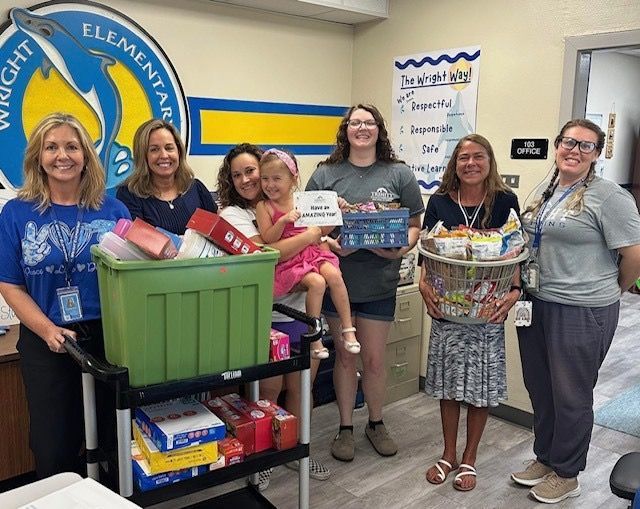 Six people in a school room stand beside a cart of donated school supplies.