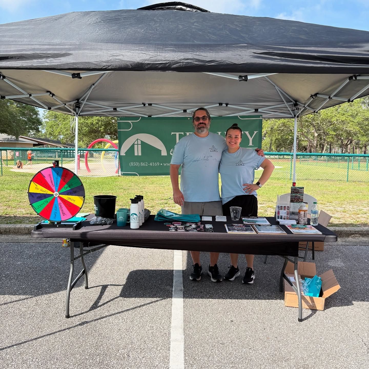 Two people under a canopy at an outdoor booth with a colorful prize wheel and table displays