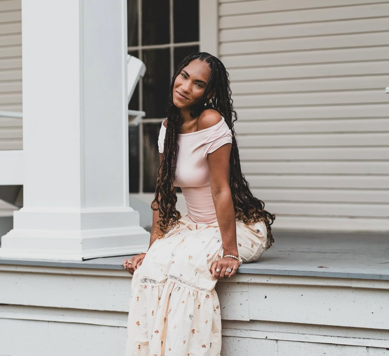 Woman in a pink top and light skirt sitting on a porch, smiling beside a white column.