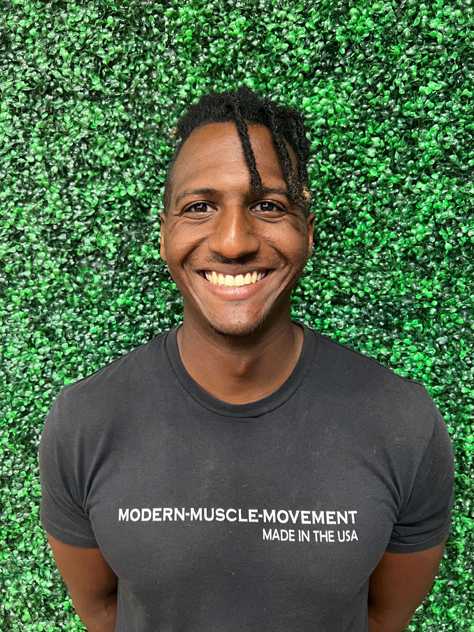 Smiling person in a dark gray T-shirt standing before a green leafy wall