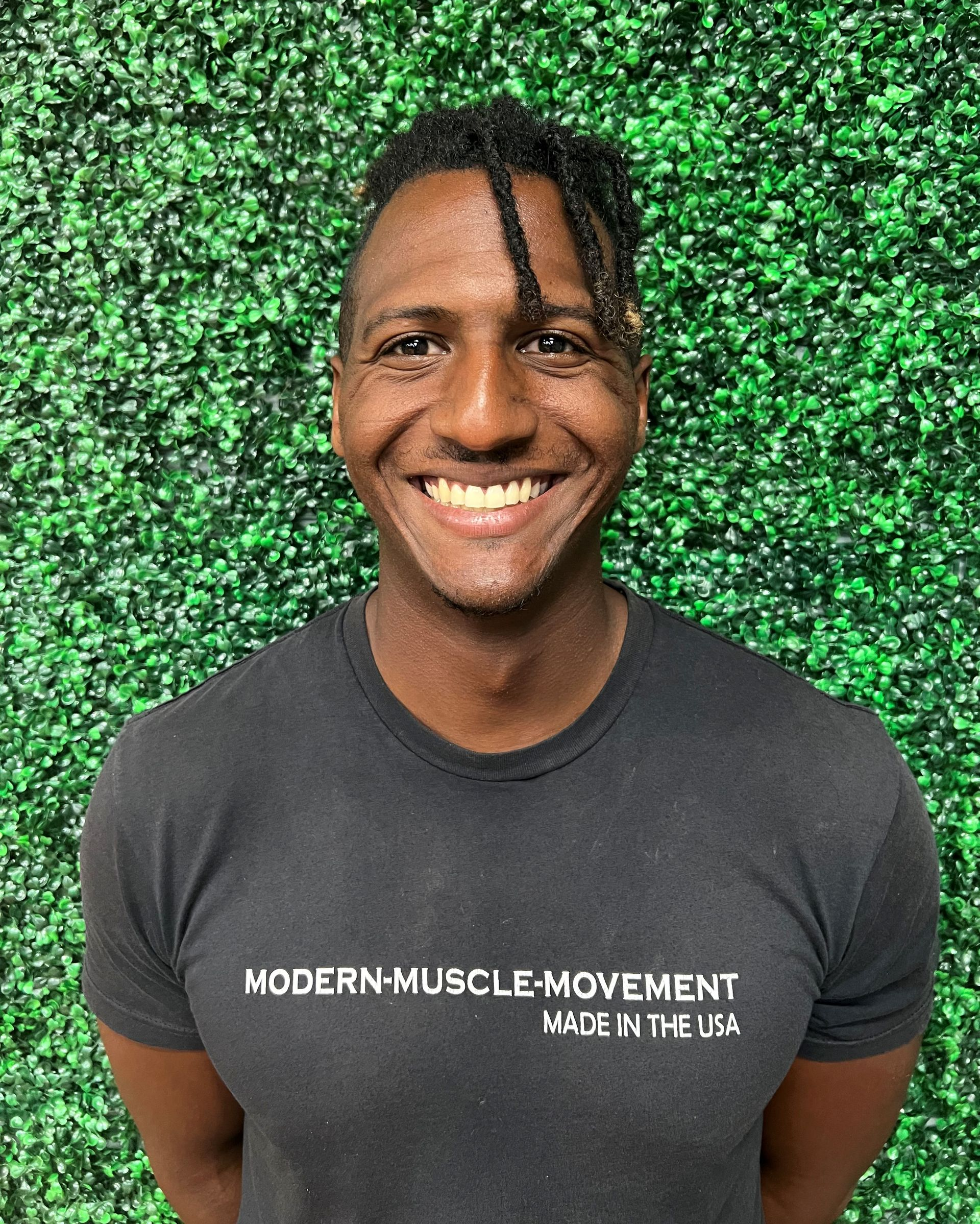 Smiling person in a dark gray T-shirt standing before a green leafy backdrop.