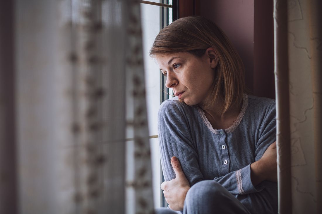 Woman sitting by a window, looking out pensively with arms crossed.