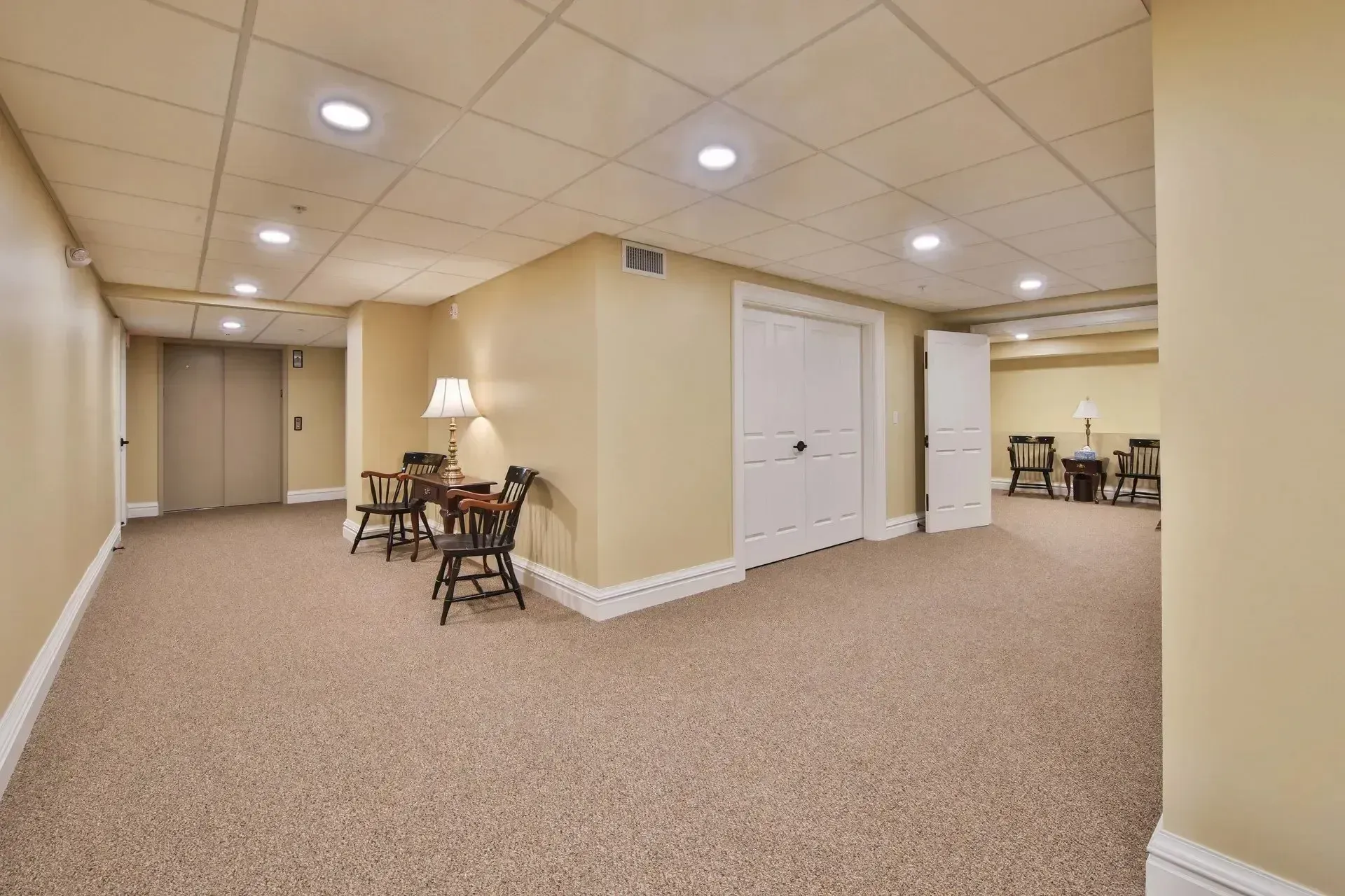 Empty beige basement room with carpet, recessed lights, a small table, and white closet doors