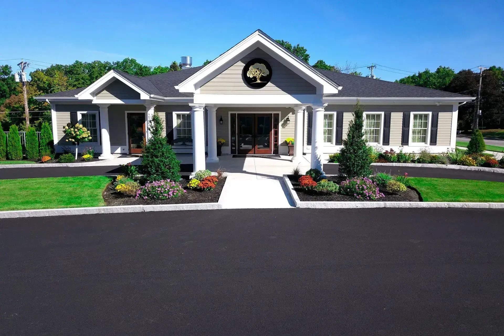 Single-story building with white columns, dark roof, landscaped entrance, and large black parking lot in front