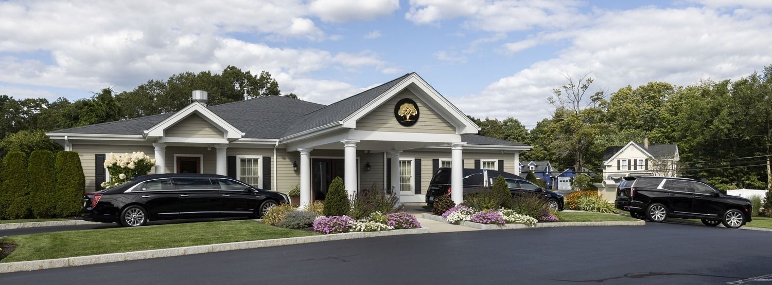 Single-story house with black roof, white columns, and flower beds beside two parked cars under a cloudy sky