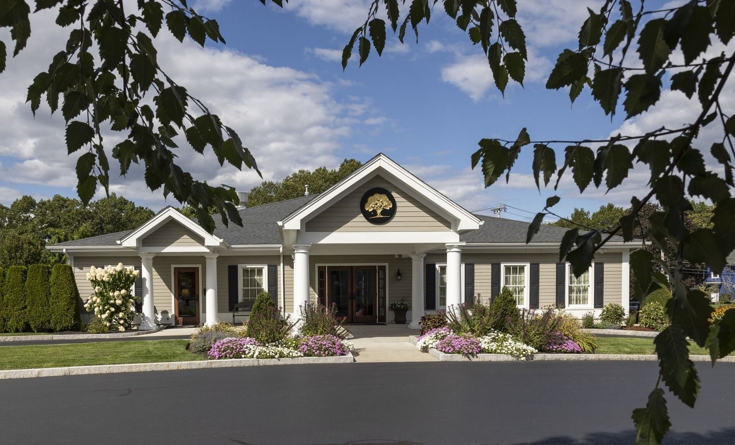 Single-story cream building with white columns and a central gable, framed by trees and flower beds
