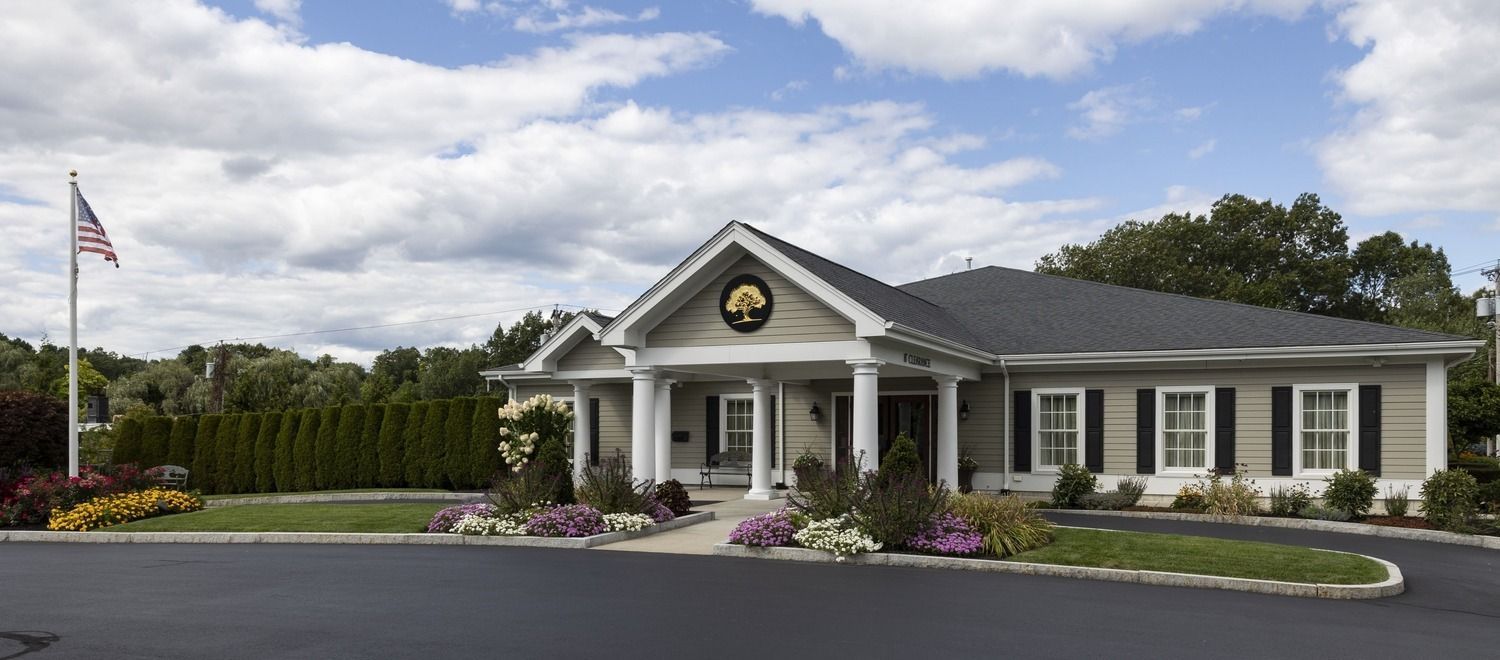 Single-story gray building with white columns, American flag, and landscaped flower beds under a cloudy sky