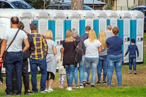 People standing in line outside blue portable toilets at an outdoor event