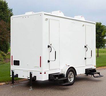 White mobile trailer with side doors, rear steps, and stabilizing jacks parked on a road.