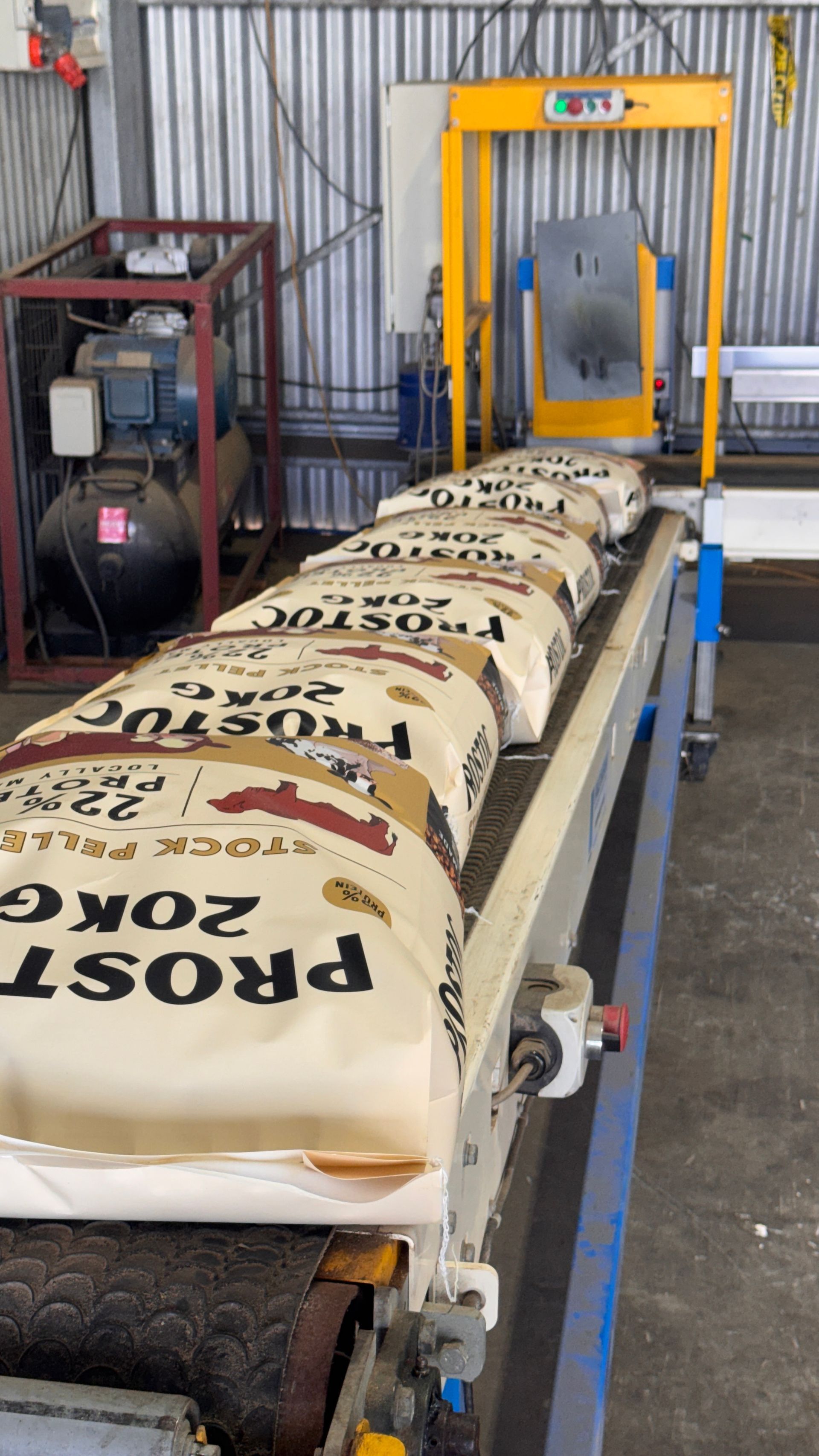 A row of labeled 20kg grain bags moves along an industrial conveyor belt toward a yellow packaging machine. — Prostoc In South Kempsey, NSW