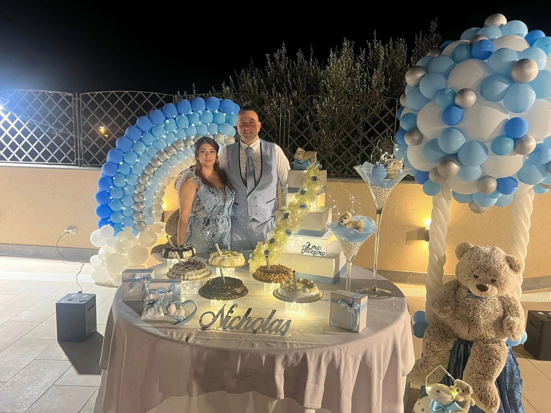 Una coppia è in piedi attorno a un tavolo decorato per un baby shower. Sono presenti palloncini blu e bianchi, dolci e un orsacchiotto di peluche.