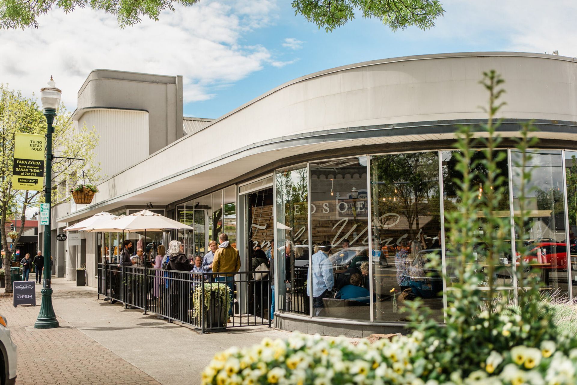 Street-side café with outdoor seating and people under umbrellas beside a curved white building.