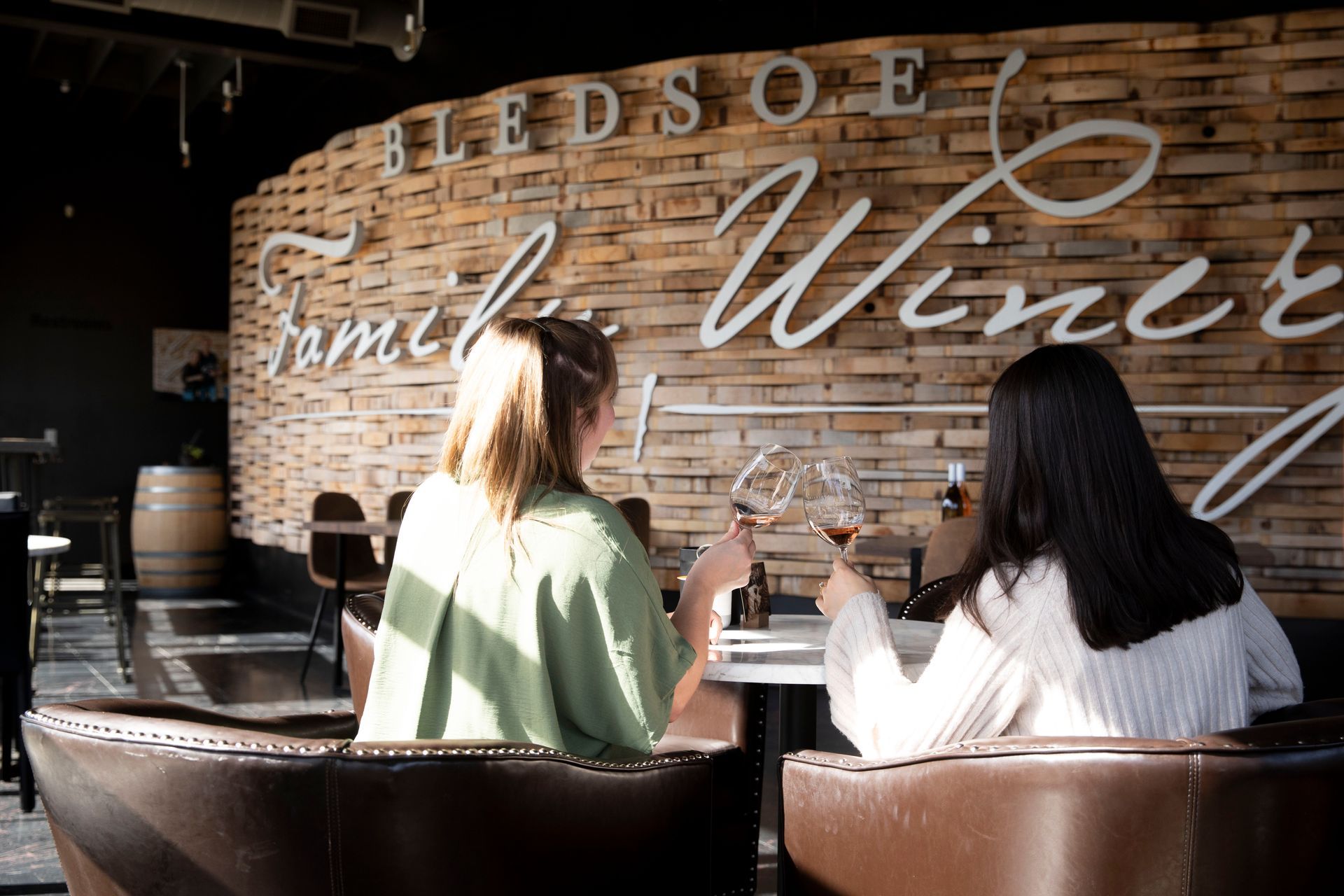 Two people seated at a winery bar, seen from behind, with a rustic wood wall reading “Winery” behind them