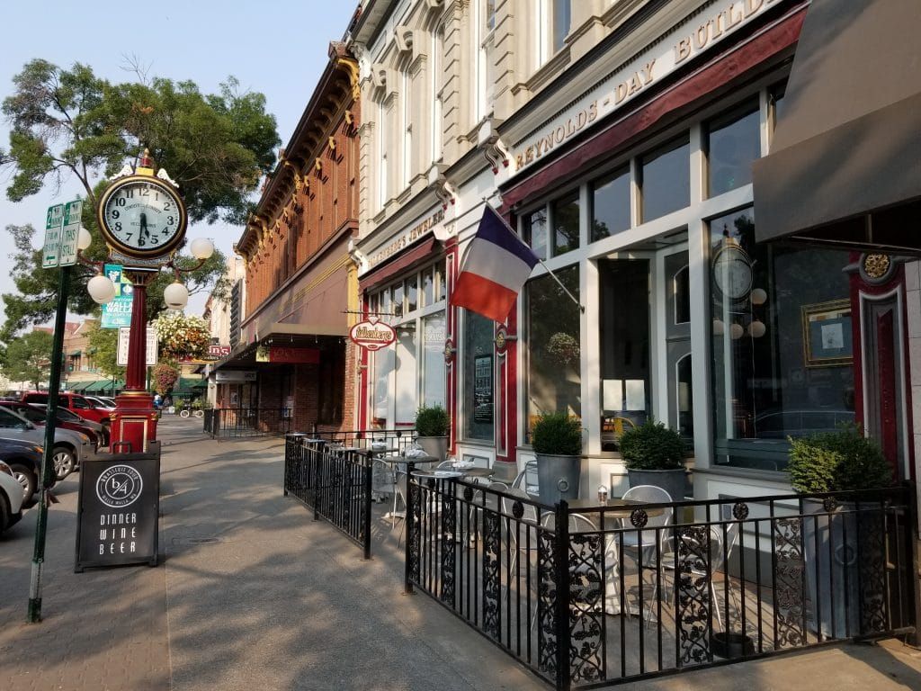 Tree-lined sidewalk with storefronts, outdoor seating, and a clock sign on a sunny street.