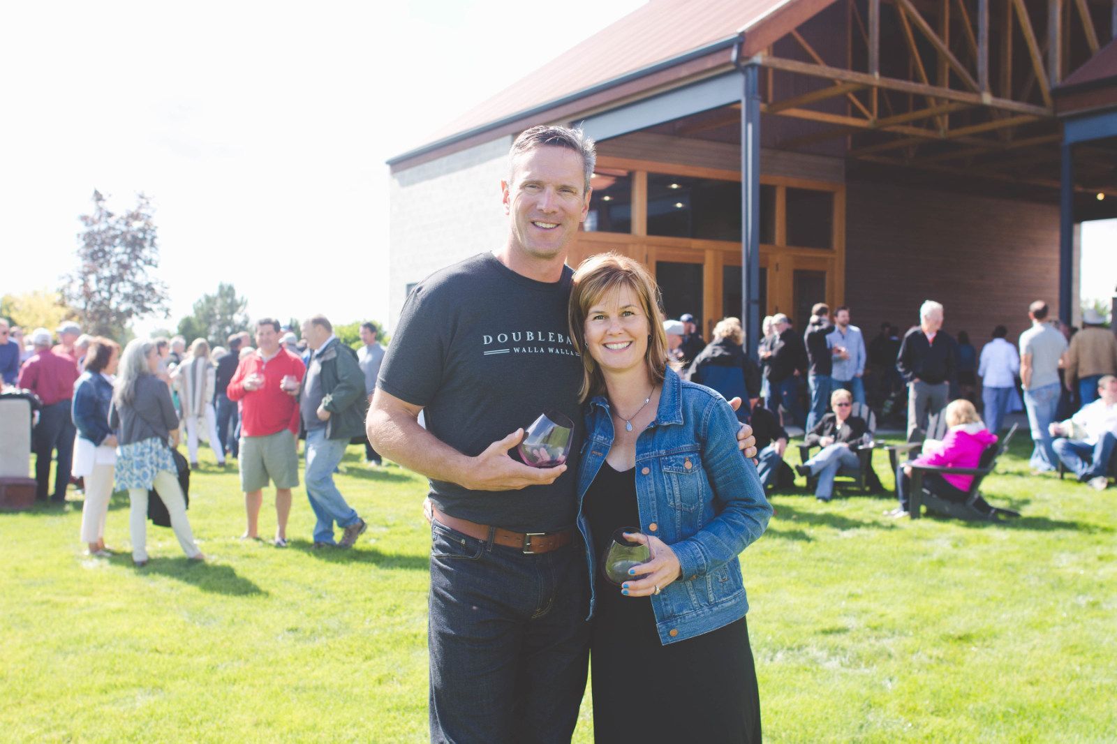 Two people smiling and holding drinks at an outdoor gathering near a barn on a sunny day