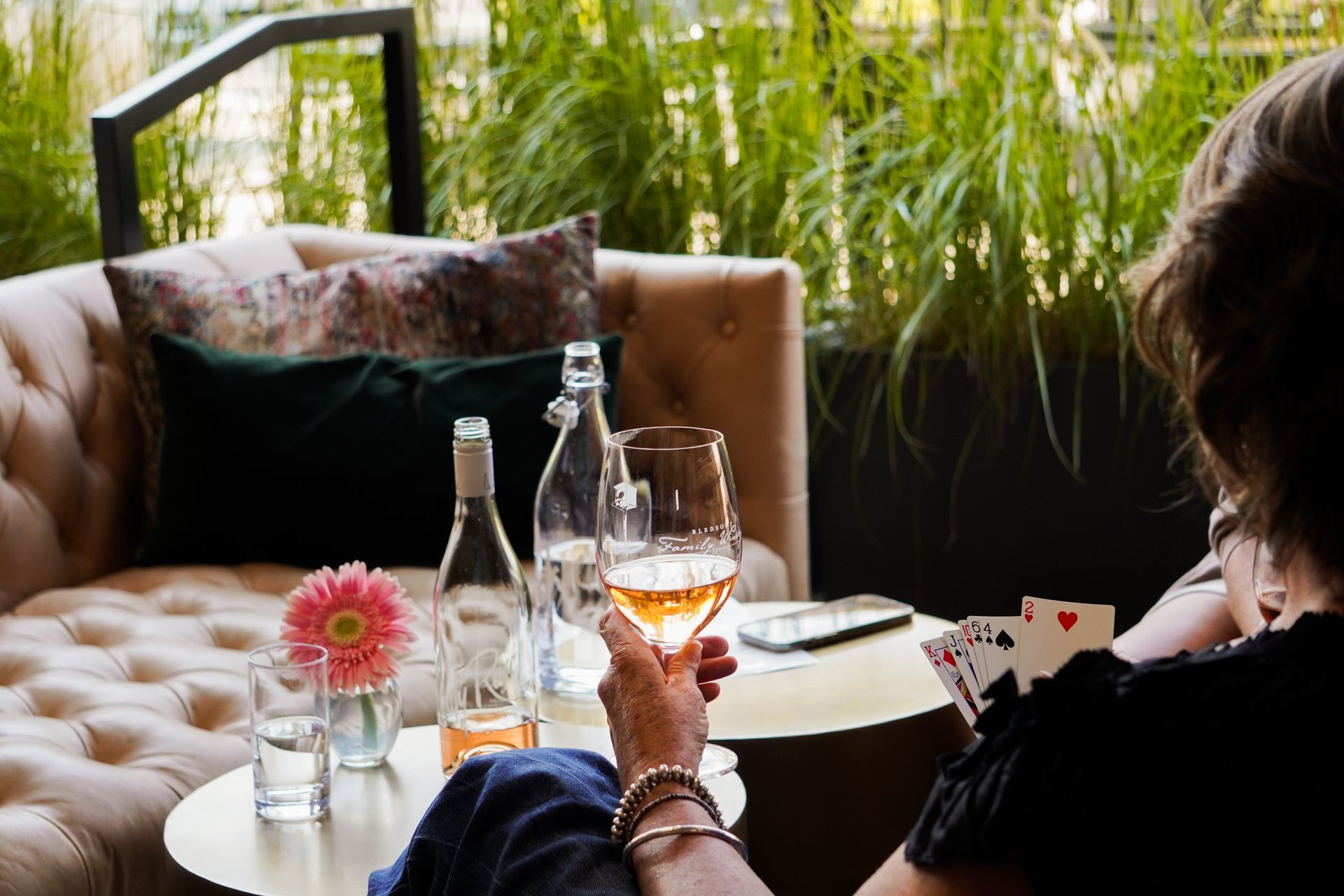 Person sipping wine at a cozy outdoor café table with flowers and playing cards