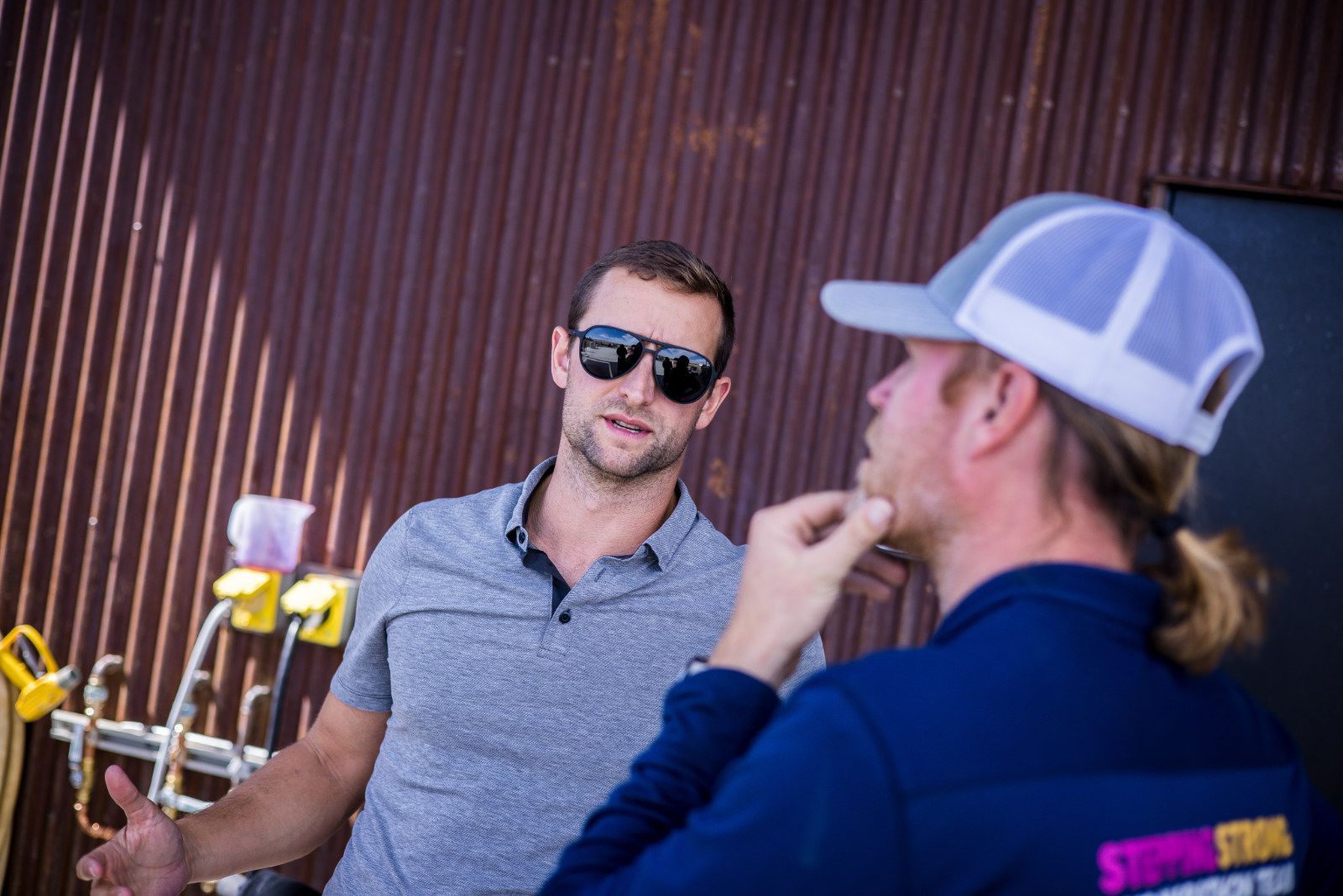 Two men talking outdoors near a wall, one wearing sunglasses and a gray shirt, the other in a white cap and blue jacket