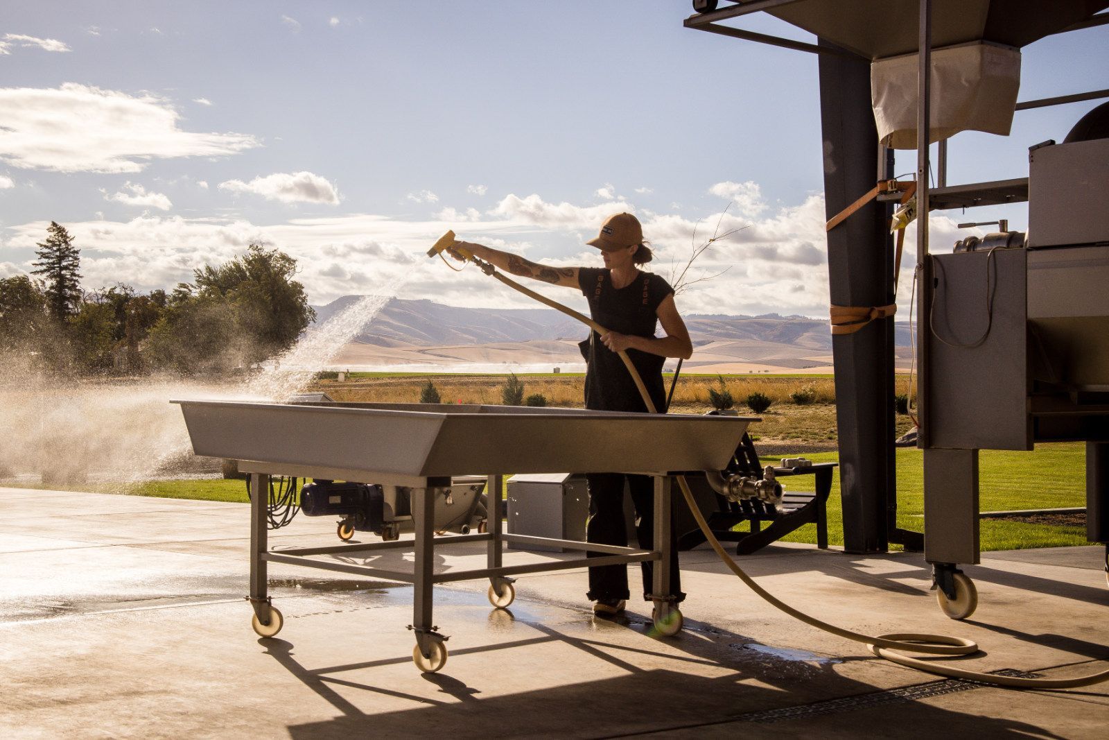 Worker spraying water on a conveyor table in a dusty outdoor industrial setting at sunset