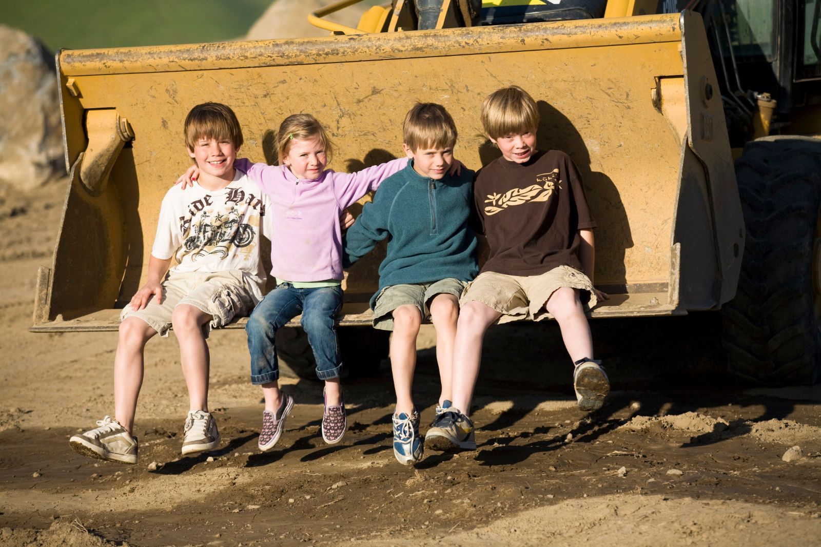 Four children sit on a large yellow construction loader bucket outdoors, smiling with dusty clothes and boots