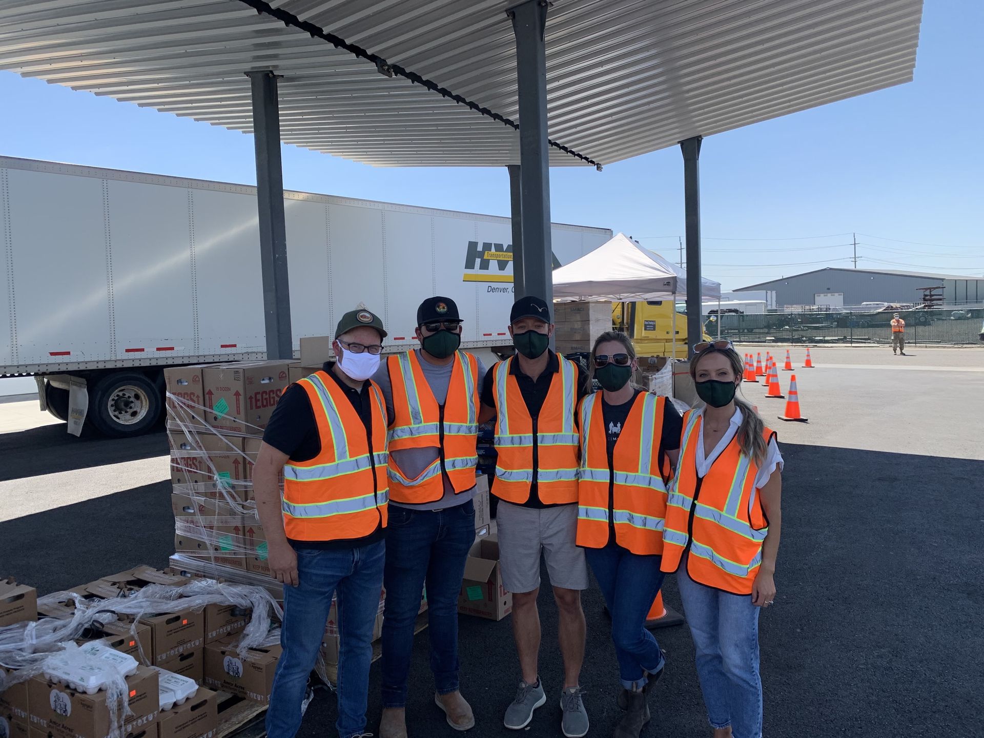 Four workers in orange safety vests and masks posing at a warehouse loading dock with pallets nearby