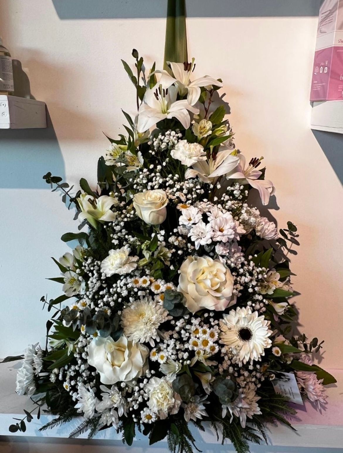 Arreglo floral blanco para funeral con rosas, margaritas y follaje en una composición triangular.