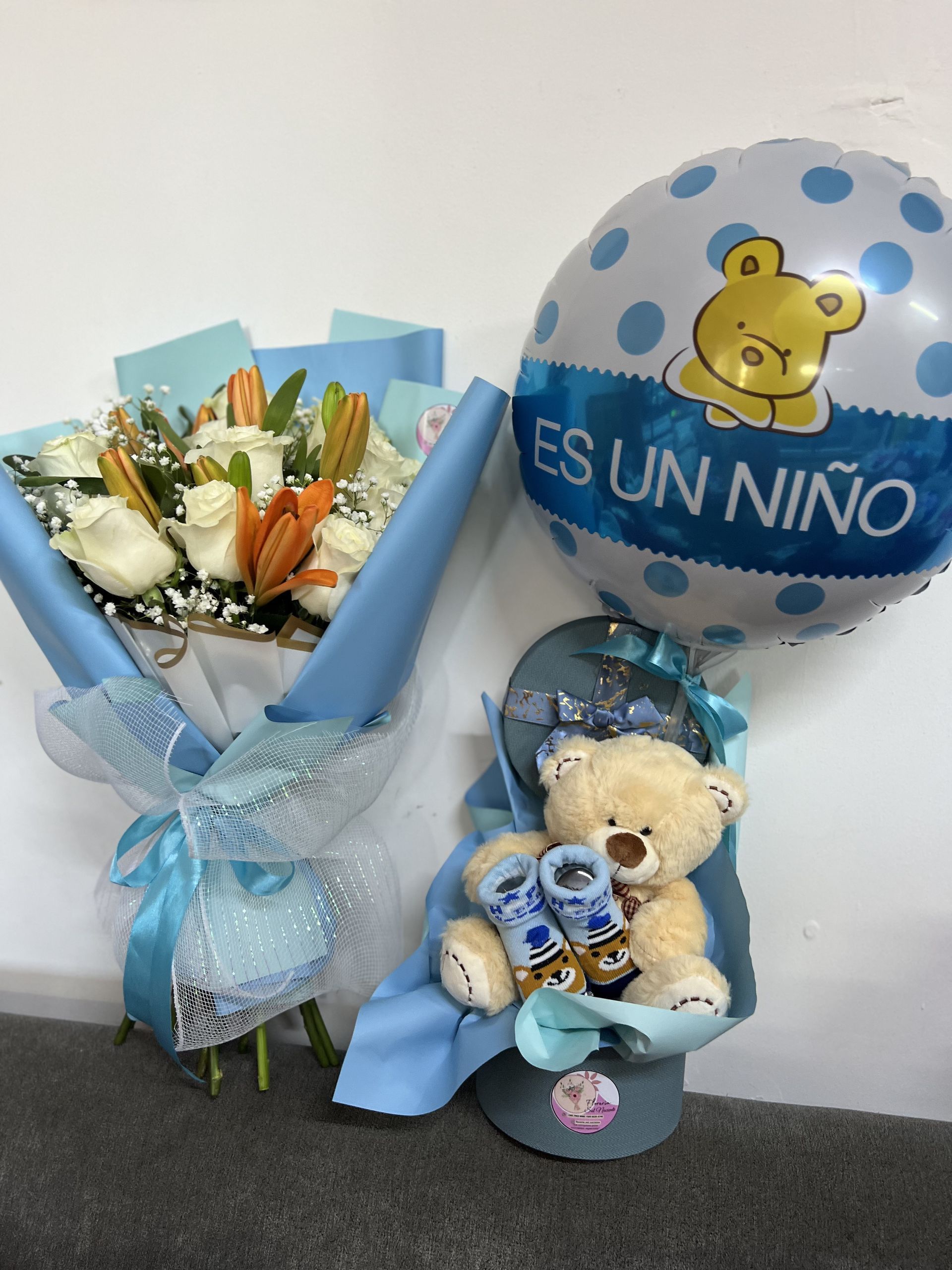 Un globo azul, una cesta de regalo con un osito de peluche y un ramo de flores blancas sobre un suelo gris.