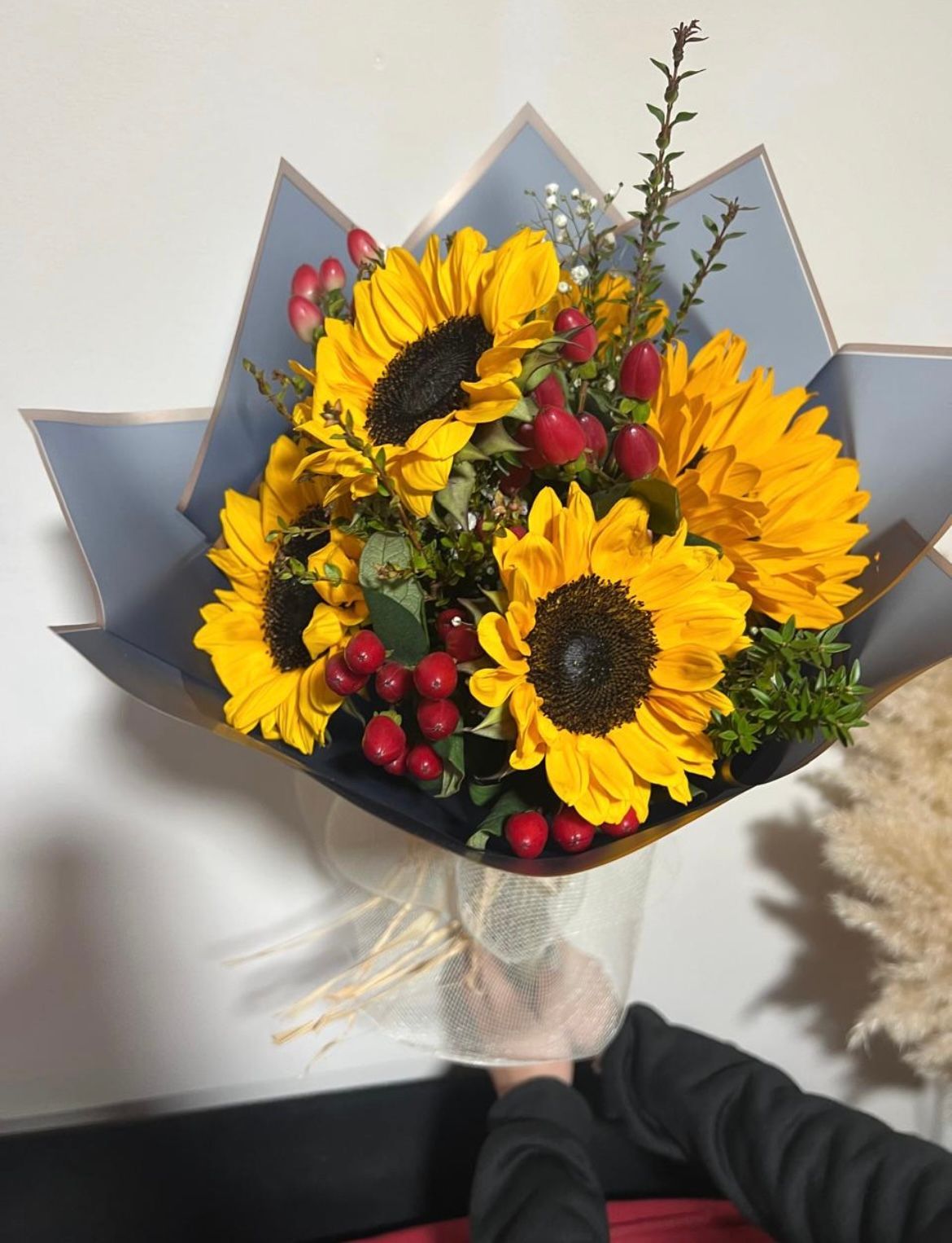 Ramo de mano compuesto por girasoles amarillos con bayas rojas, envuelto en papel oscuro.