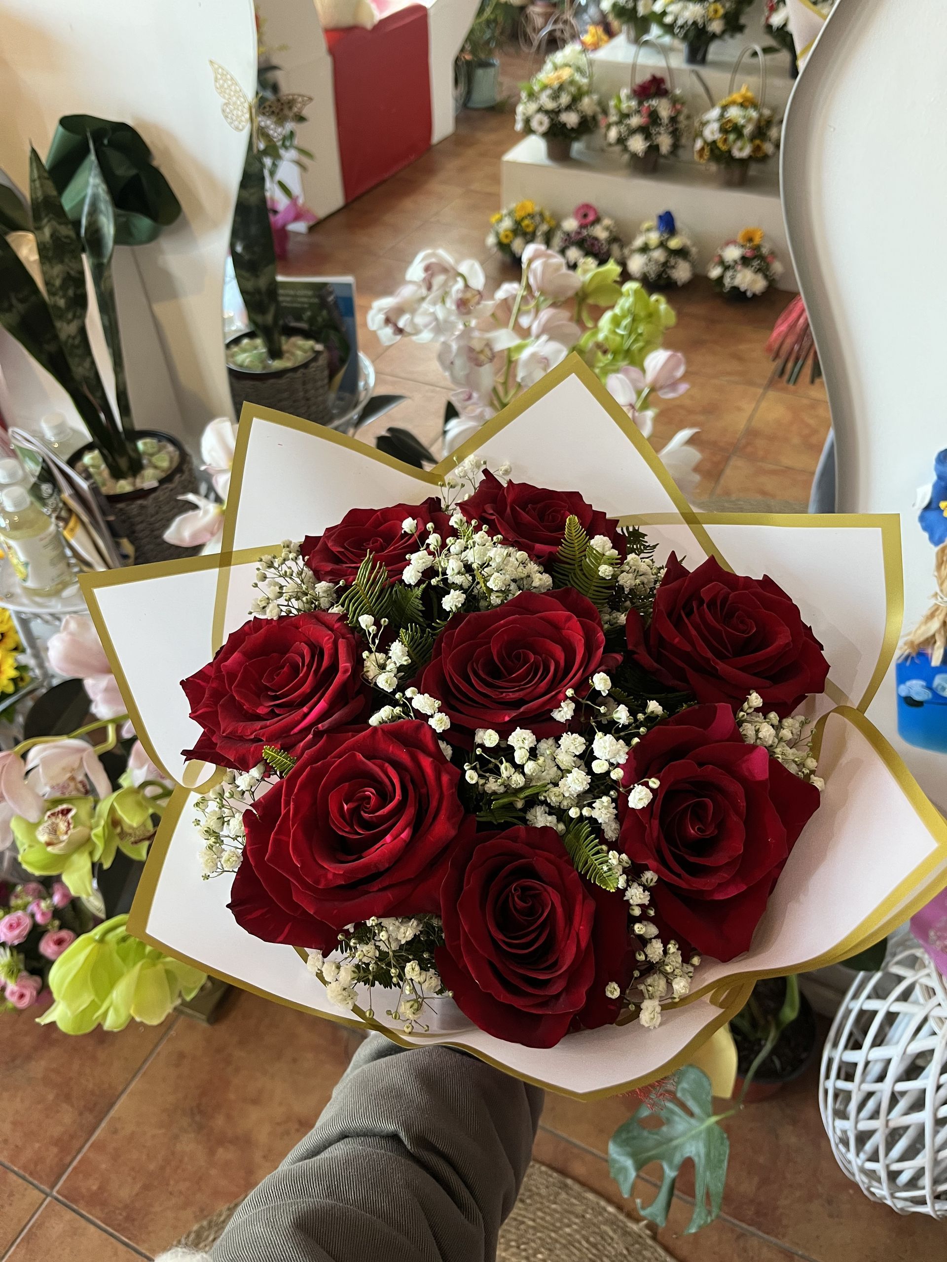 Ramo de rosas de color rojo intenso envuelto en papel blanco, sostenido en una floristería con otros ramos al fondo.