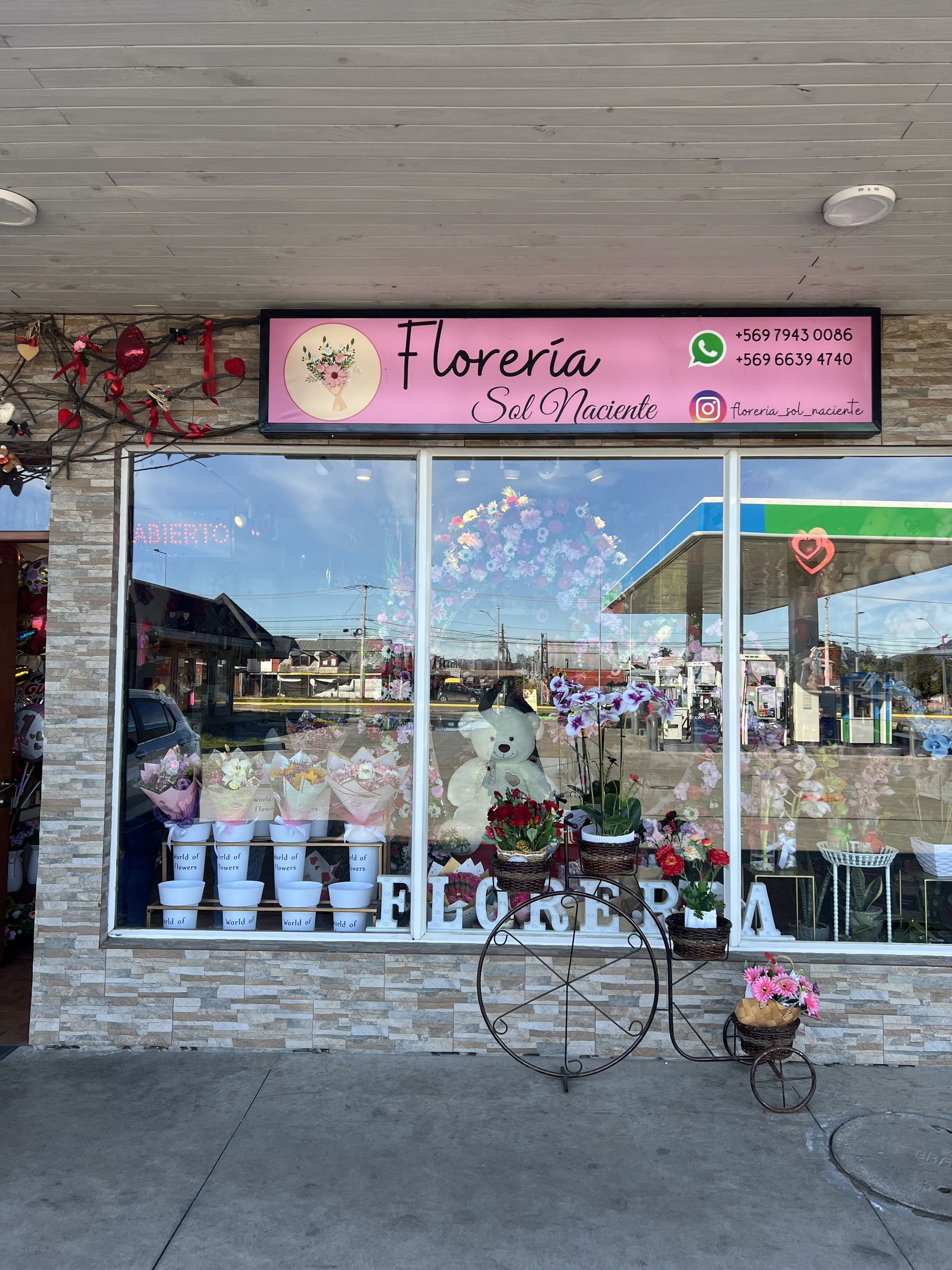 Fachada de floristería con letrero rosa, gran escaparate y puesto de flores en la acera.