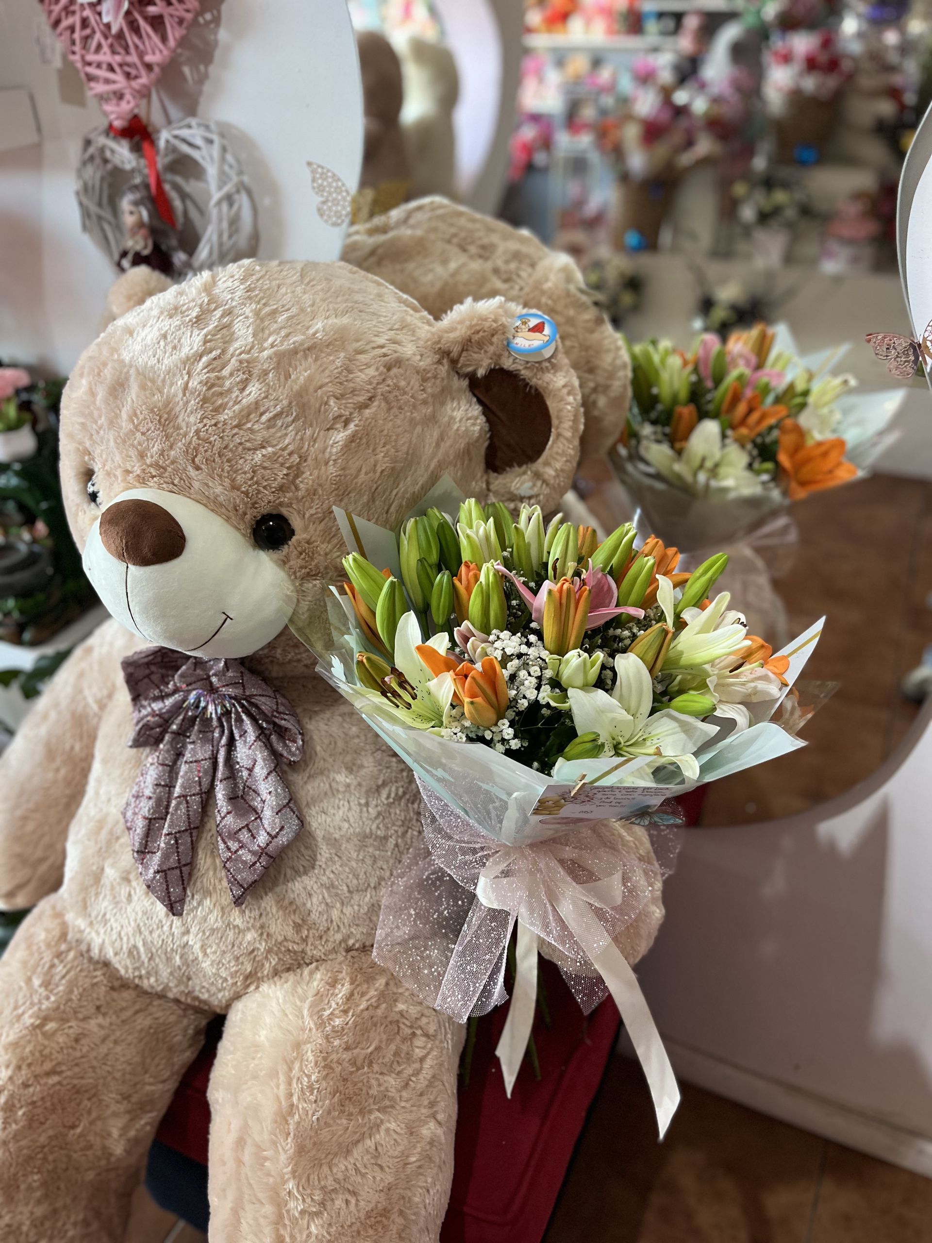 Un osito de peluche sosteniendo dos ramos de flores de colores, con una cinta morada y un fondo de papel de regalo.