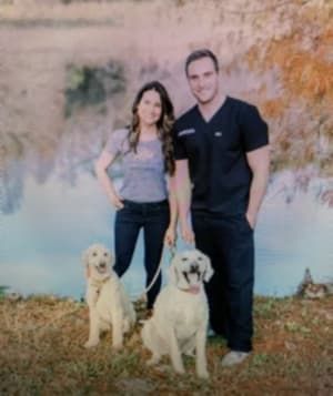 Doctor Andrew Vallo is standing in front of a pond with his wife holding their two dogs.