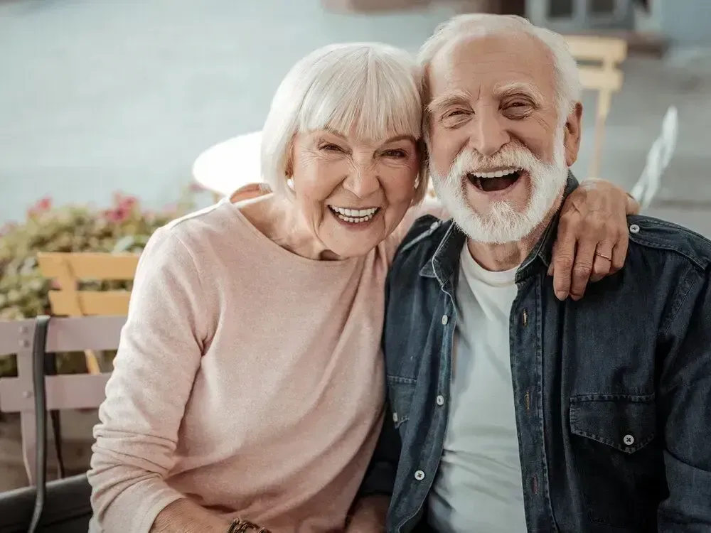 An elderly couple is sitting on a bench and smiling for the camera.