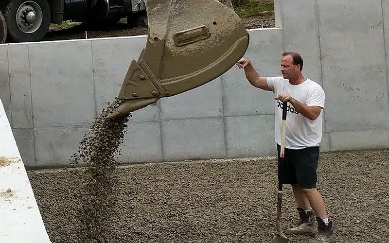 Man in white shirt guiding a concrete mixer chute as wet concrete pours onto the ground indoors