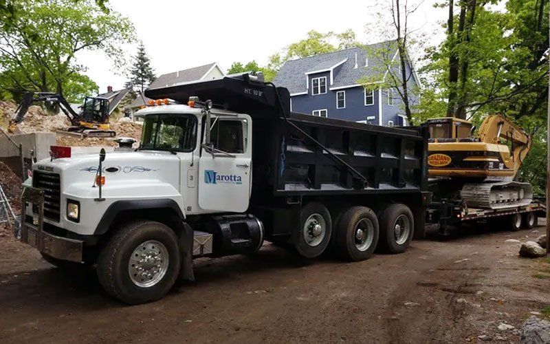 White dump truck hauling a yellow skid steer on a lowboy trailer along a muddy residential street