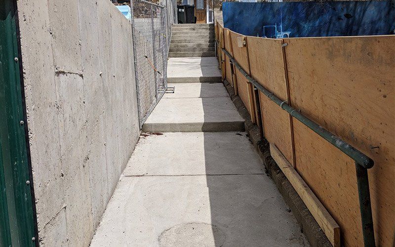 Narrow concrete walkway between high walls, with a railing on the right and steps ahead in bright sunlight