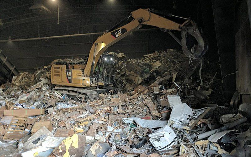 Yellow excavator clearing a large pile of debris inside a dark warehouse
