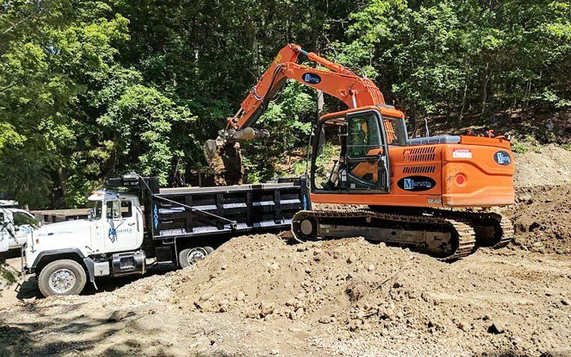 Orange excavator loading dirt into a white dump truck at a construction site near trees